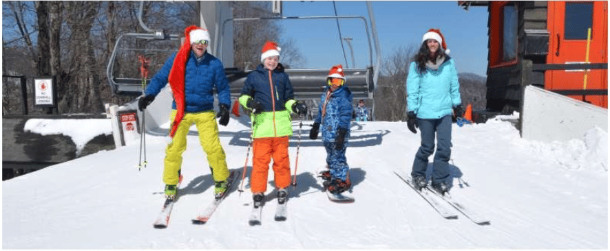 A family in Santa hats skis together on a snowy slope beside a chairlift.