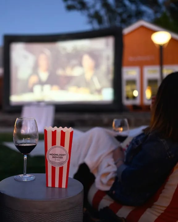 A person relaxes outdoors at dusk, watching a movie projected on a large inflatable screen. In the foreground, a glass of red wine and a red and white striped popcorn box with 
