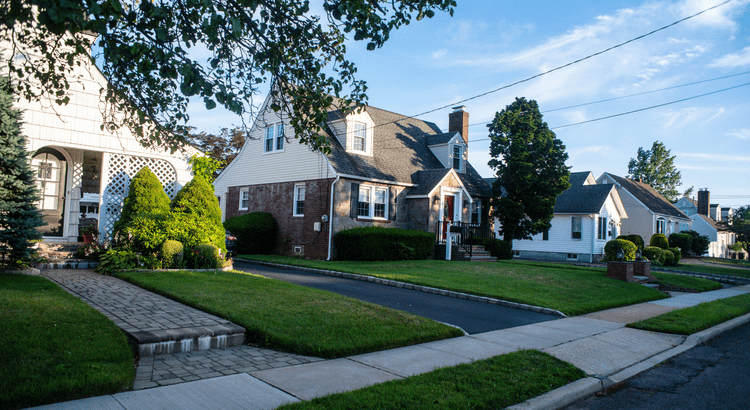 Suburban neighborhood scene with charming, well-kept houses featuring manicured lawns and trees lining the sidewalk under a clear blue sky.