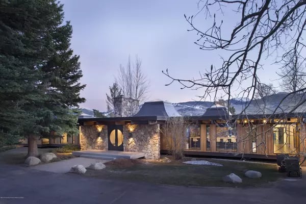 Modern stone-and-glass home with a round front door, photographed at dusk against snow-dusted mountains.