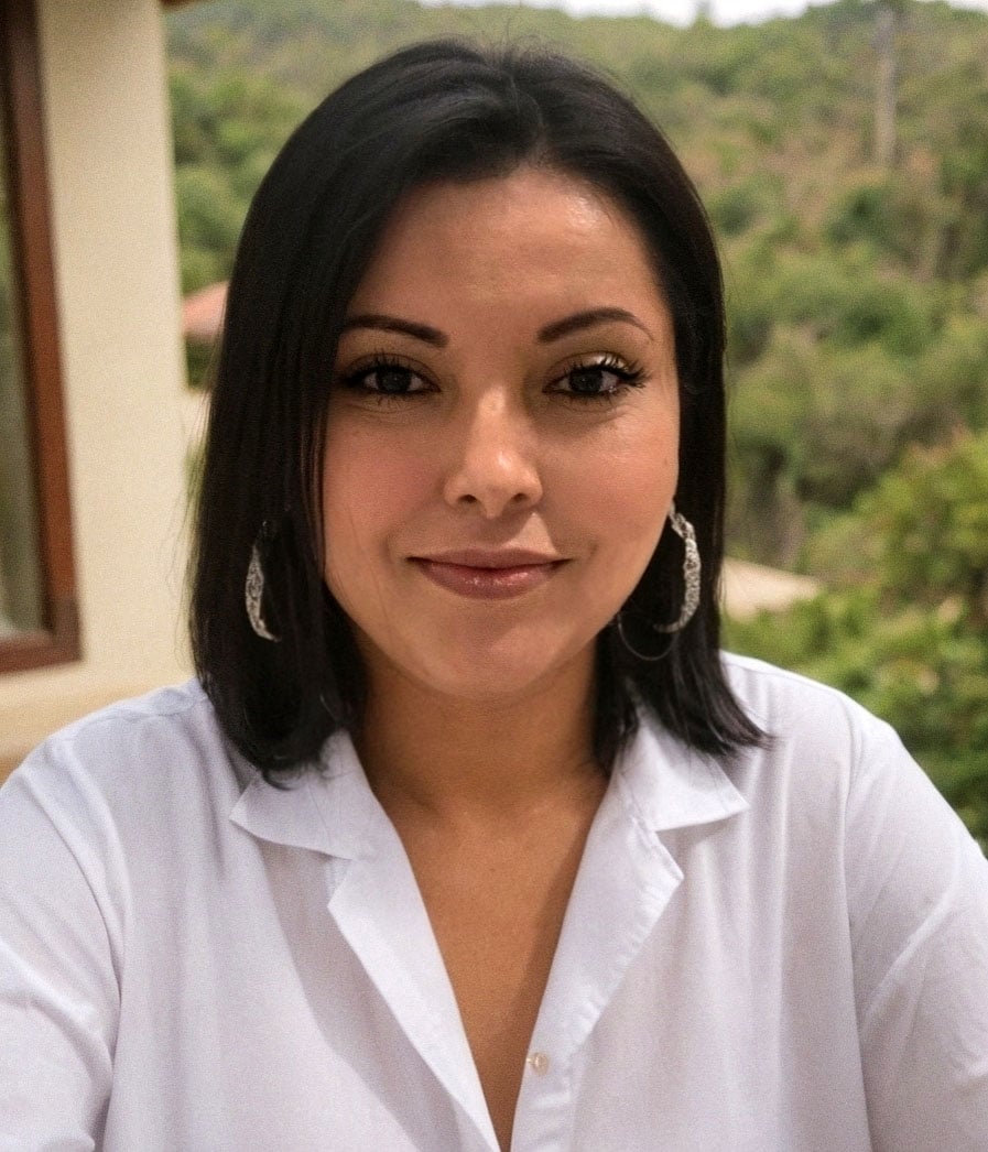 Victoria Tschirhart in a white blouse and hoop earrings, with greenery behind her.