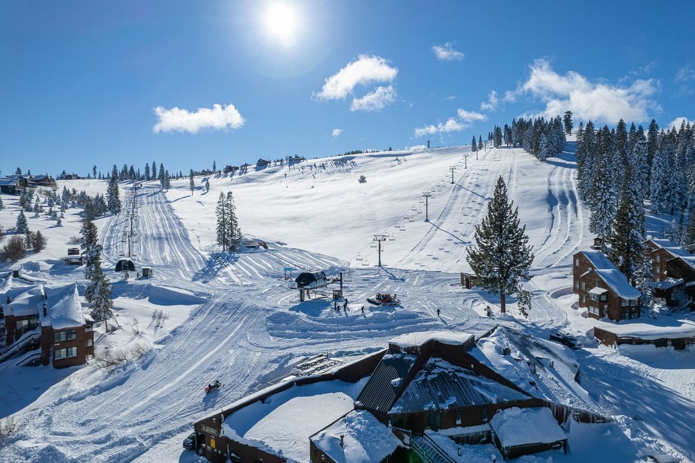 Snow-covered ski lodge below groomed ski runs and chairlifts on a sunny winter day.