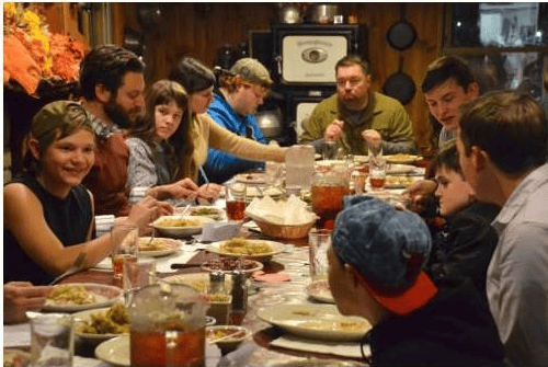 Family and friends share Thanksgiving dinner around a crowded table in a wood-paneled room.