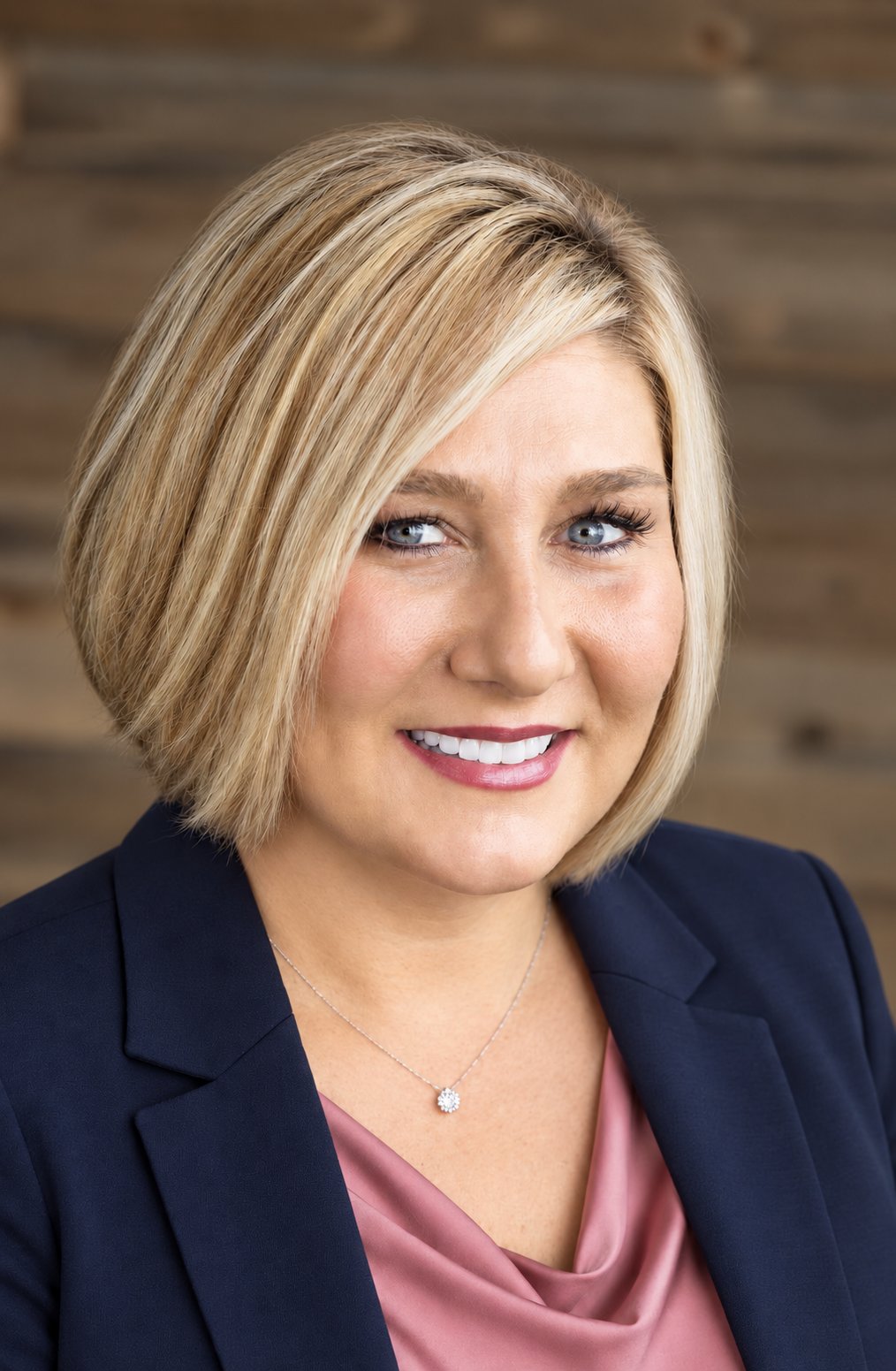Smiling blonde woman in a navy blazer and pink blouse, wearing a small pendant necklace.