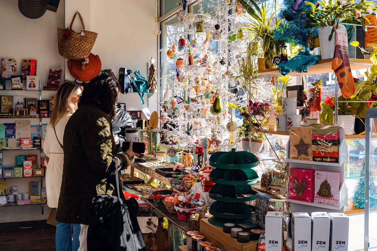 Shoppers browse a sunlit gift shop filled with colorful ornaments, cards, plants, and holiday decorations.