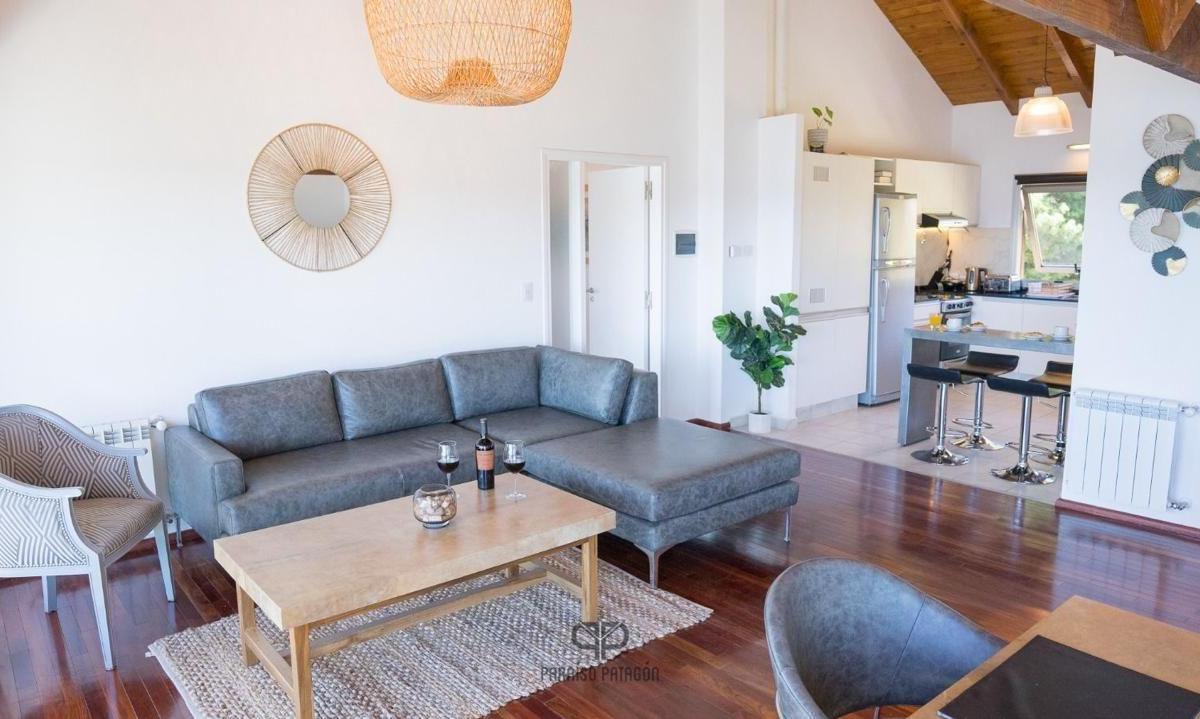 Living room with a gray sectional, stone coffee table, and open kitchen beneath a vaulted wood ceiling.