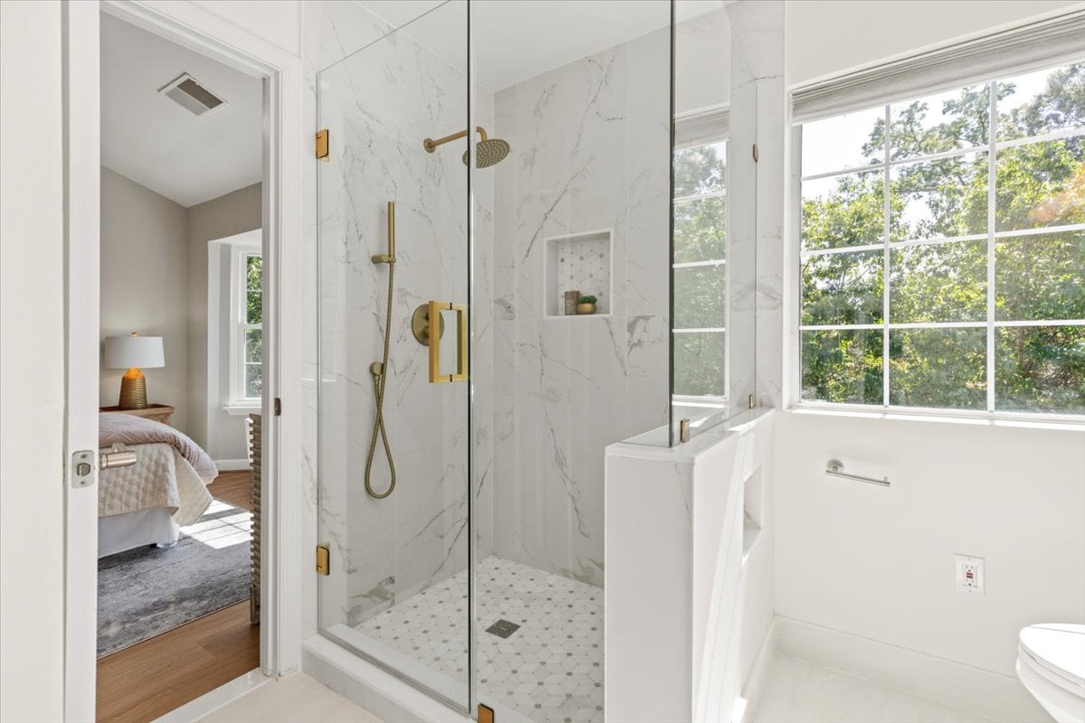 Bright bathroom with a marble-look glass shower, brass fixtures, and large windows overlooking trees.