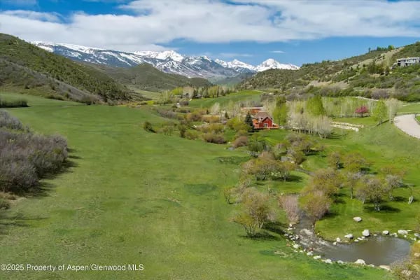 Aerial view of a green mountain valley with homes, a creek, and snowy peaks.