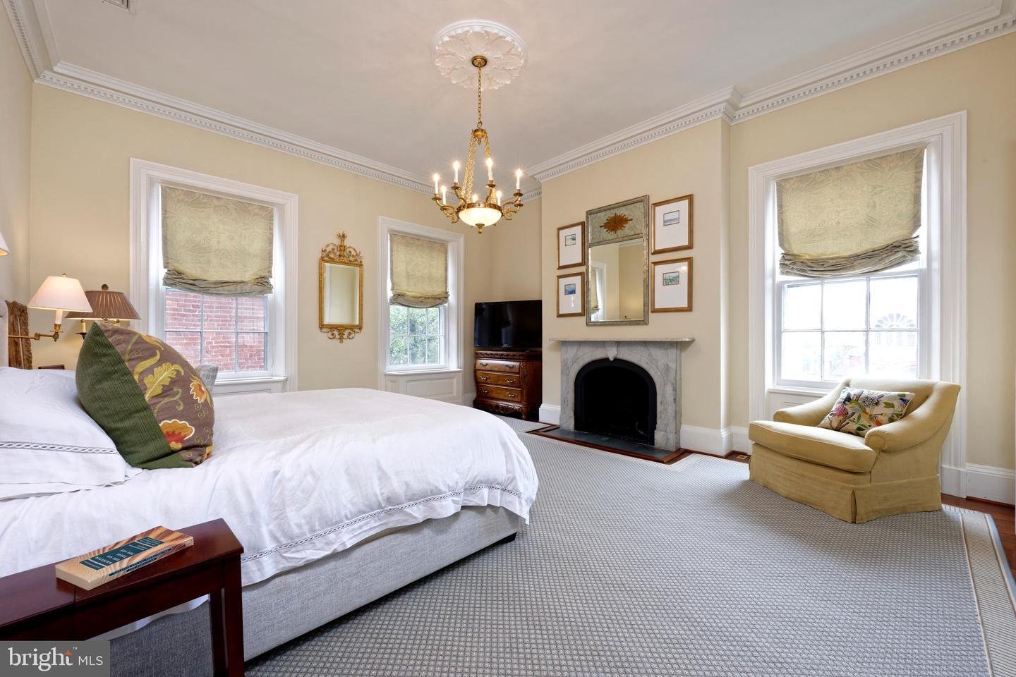 Bedroom with a large white bed, marble fireplace, ornate chandelier, and armchair by tall windows.