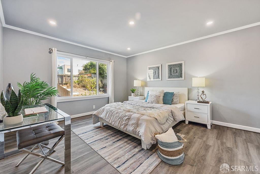 Staged bedroom with a large window, upholstered bed, mirrored desk, and gray wood flooring.