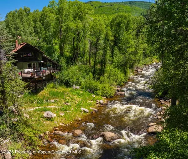 Wooded mountain cabin beside a rocky creek, with a raised deck and lush summer foliage.
