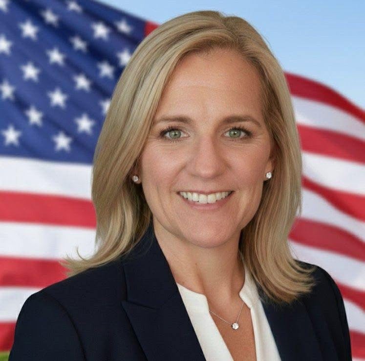 Kandice Hill smiling in a navy blazer with an American flag behind her.