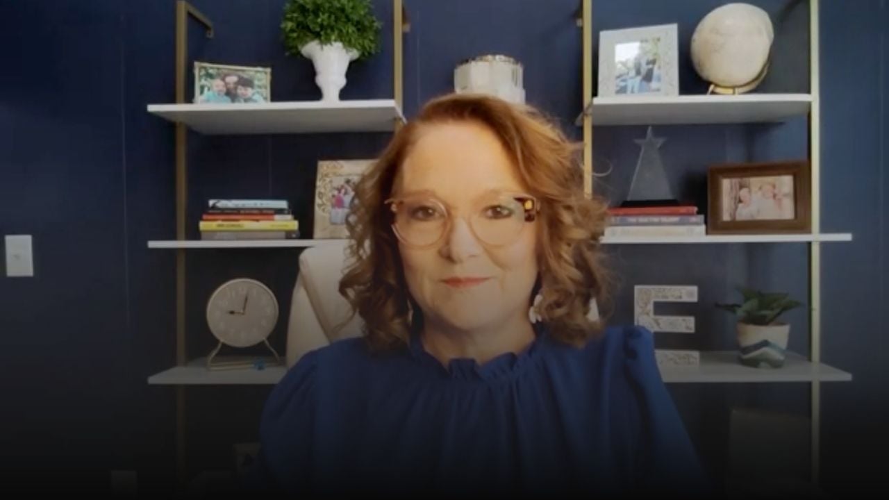 Woman with glasses speaking to camera in front of dark blue office shelves.