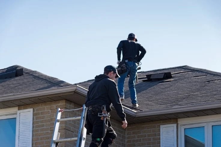 Roofers work on a brick house’s shingle roof, with one climbing a ladder and another near a roof vent.