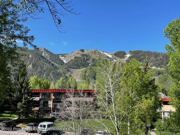 Aspen building among leafy trees, with snow-patched mountains behind it.