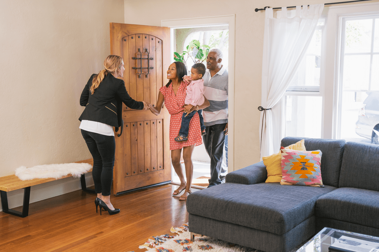 A family of three is greeted by a real estate agent in a cozy living room. The scene conveys warmth and welcome, with a couch and bright cushions.