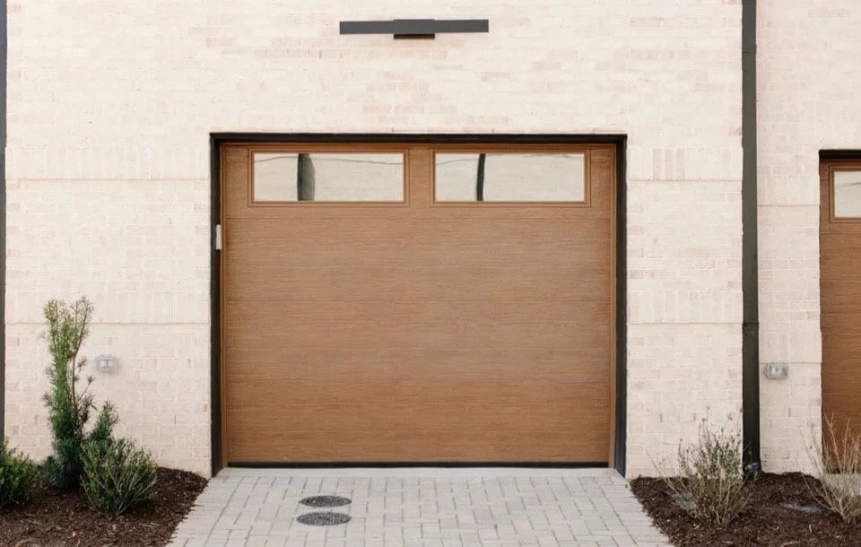 Wood-toned garage door with two narrow windows in a pale brick facade.