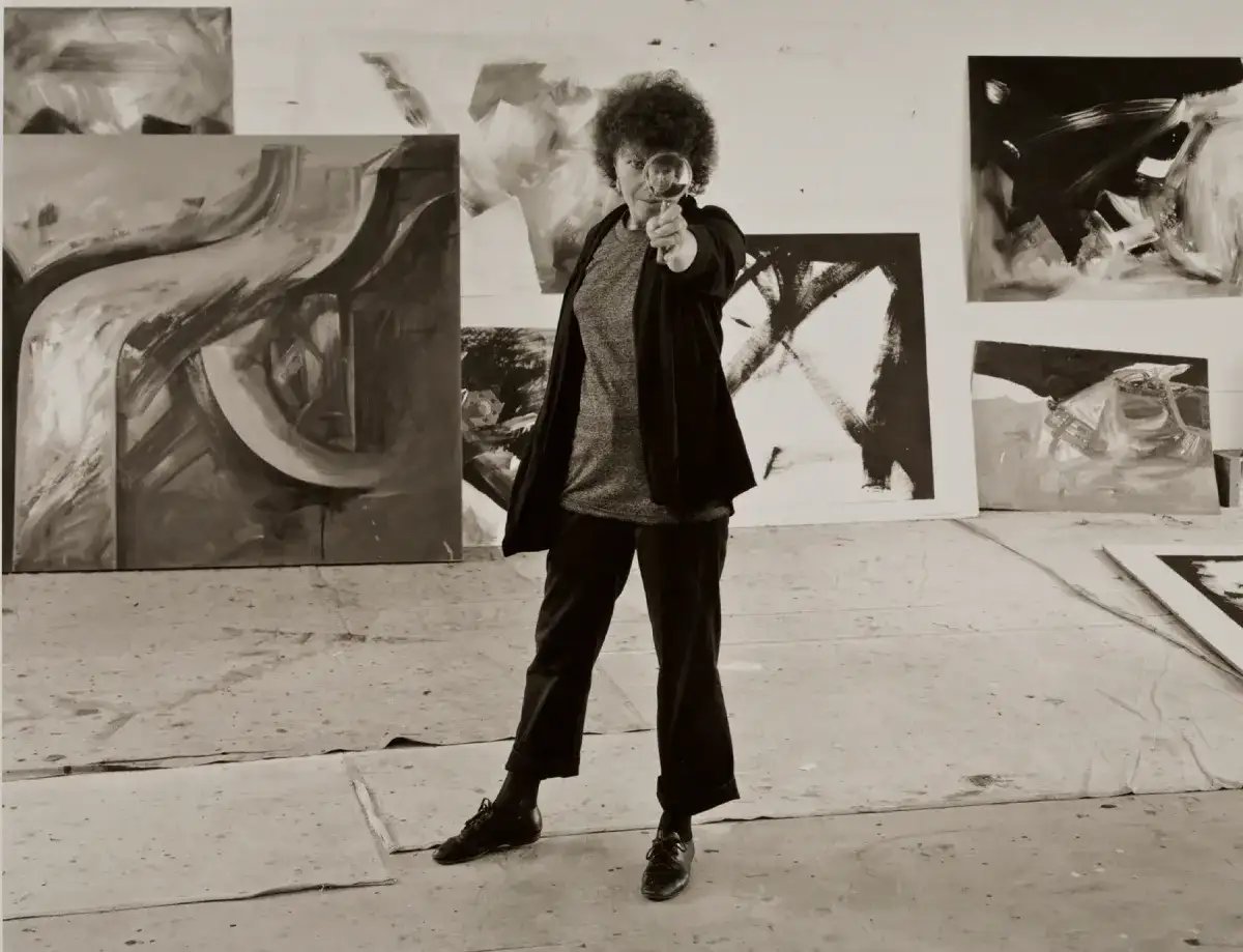 A black-and-white photo of artist Jay DeFeo in her studio, holding a magnifying glass to one eye. She wears a dark coat and trousers, standing on a concrete floor with drop cloths. Large abstract paintings with bold brushstrokes adorn the walls.