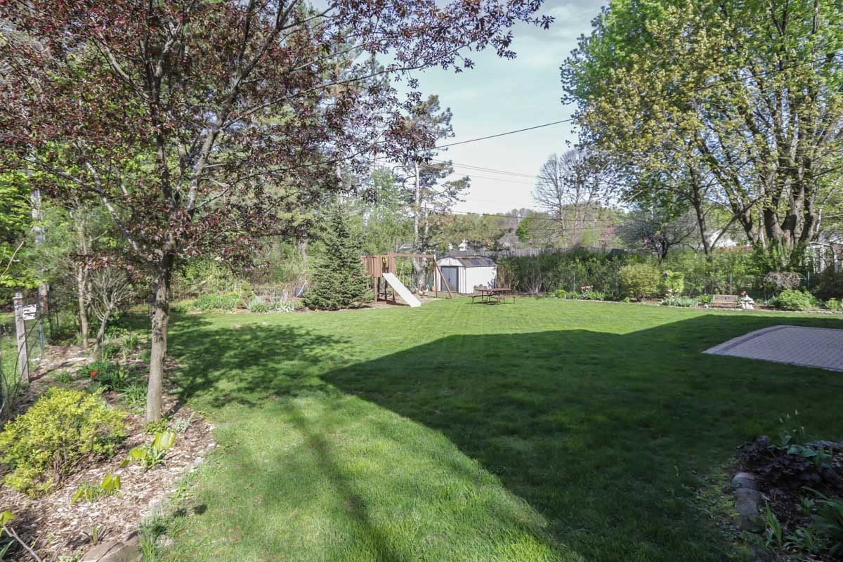 Landscaped backyard with a broad lawn, mature trees, playset, shed, and paved patio.