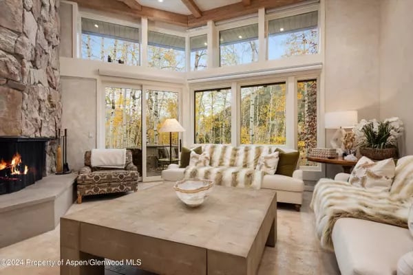 Living room with a stone fireplace, pale sofas, and tall windows overlooking autumn trees.