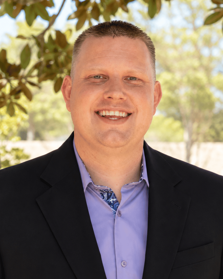 Kyle White in a black blazer and lavender shirt, smiling outdoors.