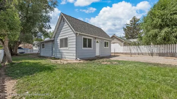 Gray detached house with a steep gabled roof, grassy yard, and white fence.