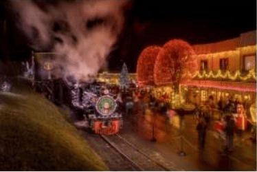 A decorated holiday train passes illuminated buildings at night.