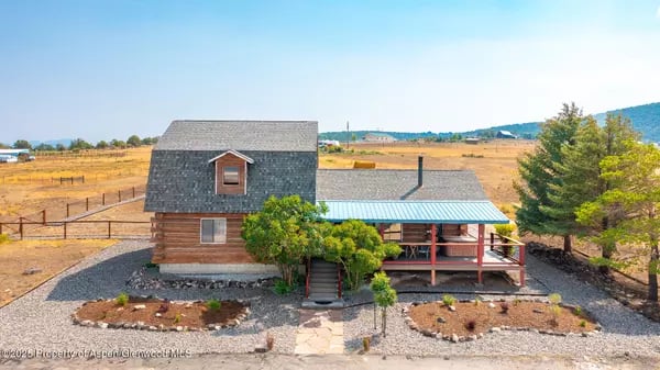 Rustic two-story home with a blue metal porch roof in an open rural landscape.