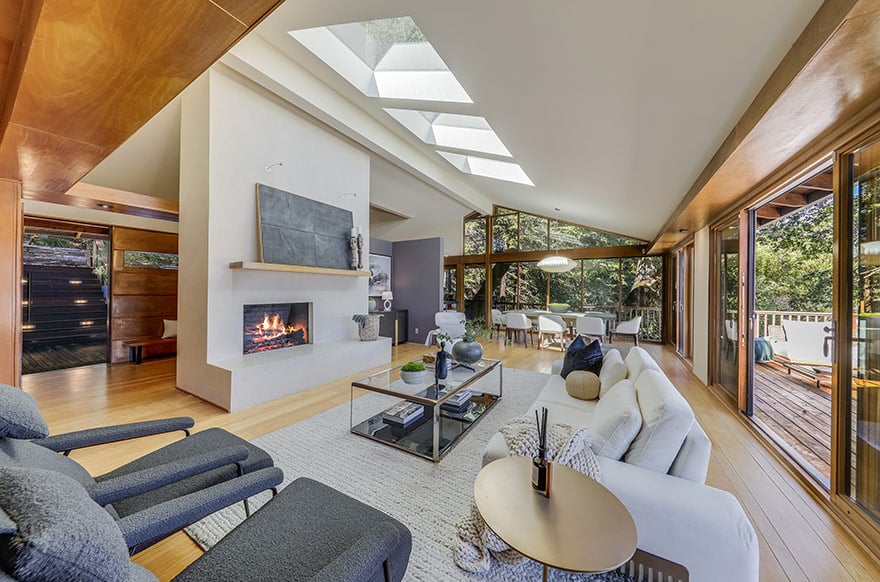 Staged living room with a fireplace, vaulted skylit ceiling, and windows overlooking a wooded deck.