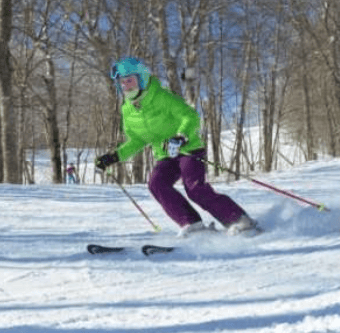 Skier carving through snow on a wooded slope, wearing a bright green jacket and purple pants.