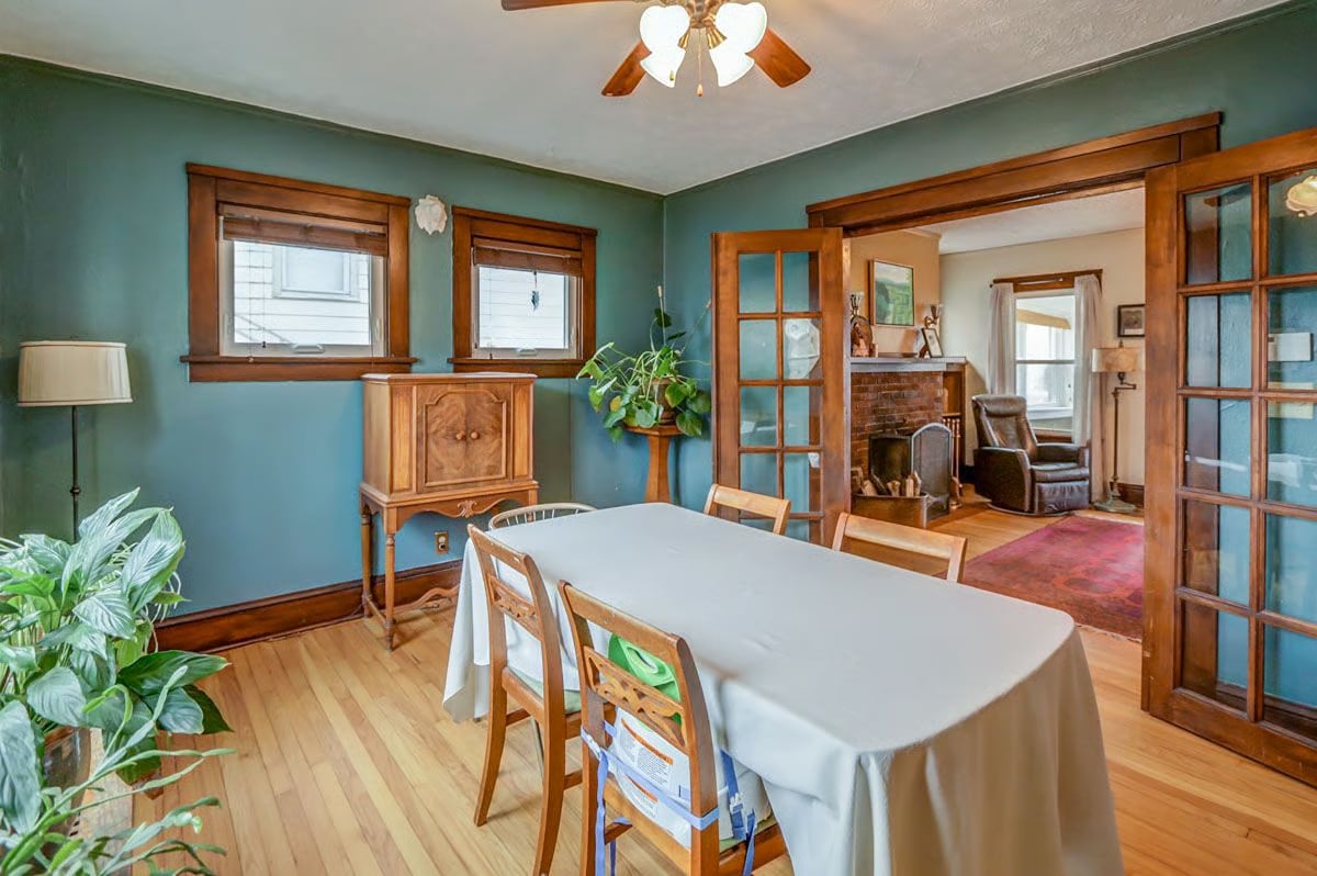 Dining room with teal walls, wood trim, a white-clothed table, and glass doors opening to a fireplace room.