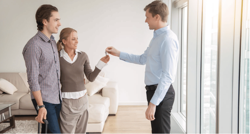 A man hands keys to a smiling woman beside another man in a bright living room.