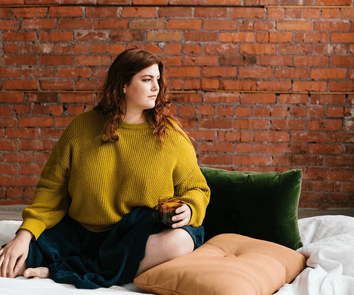 A woman in a mustard-yellow sweater holding a mug sits against an exposed brick wall.