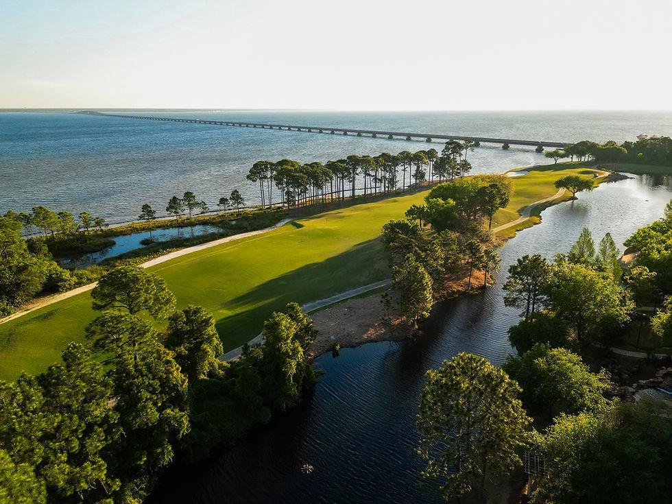 Aerial view of a waterfront golf course with tree-lined fairways and a long bridge across the bay.