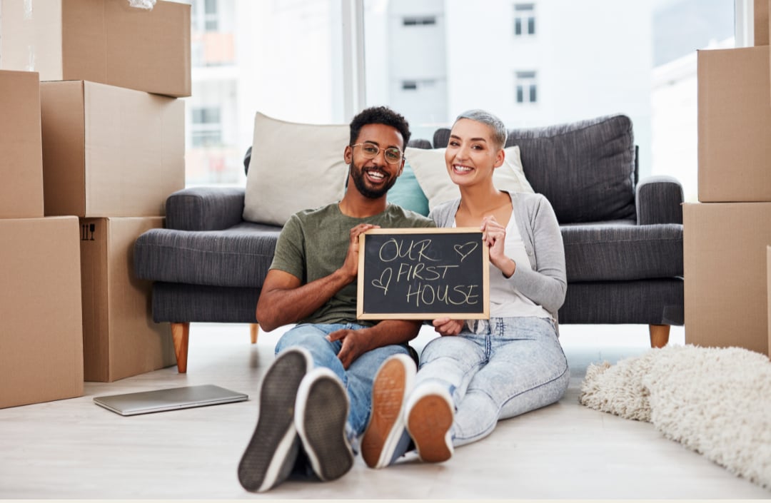 A smiling couple celebrates their first house among moving boxes, holding a sign that reads “OUR FIRST HOUSE.”