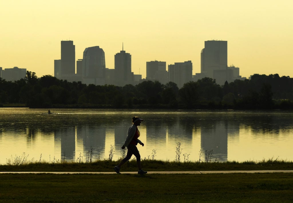 Denver skyline reflected in a lake as a person walks along the shore at golden hour.