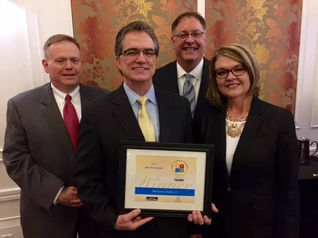 Four business professionals pose with a framed award at an indoor event.