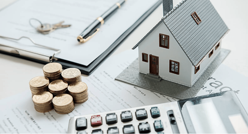 Miniature house model beside stacks of coins, a calculator, and real estate paperwork.
