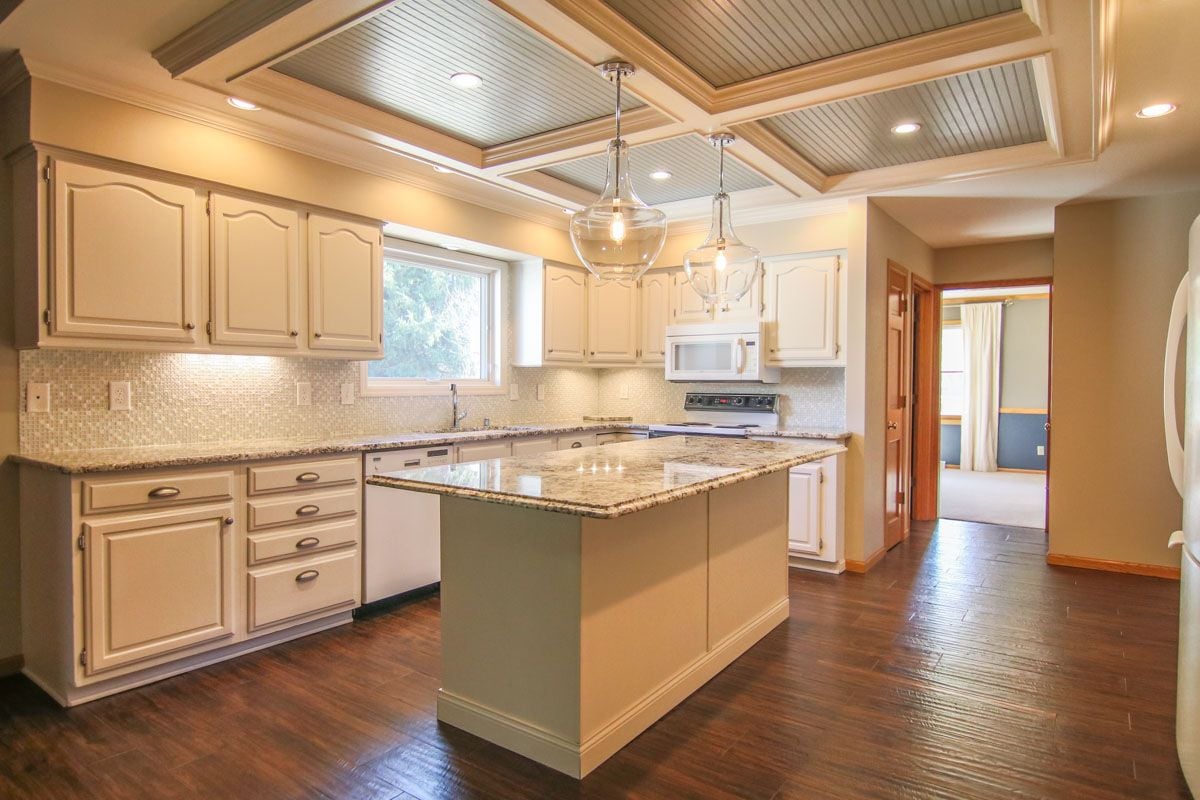 Kitchen with a granite-topped island, cream cabinetry, dark wood floors, and a coffered ceiling.
