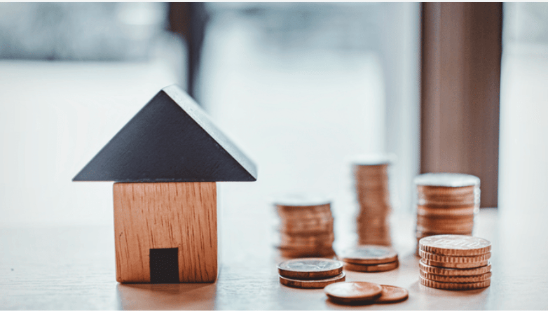 Wooden house model beside stacks of coins on a tabletop.
