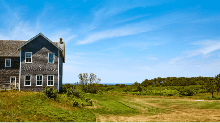 Shingle-sided coastal home beside a grassy lawn, with ocean visible beyond the dunes.