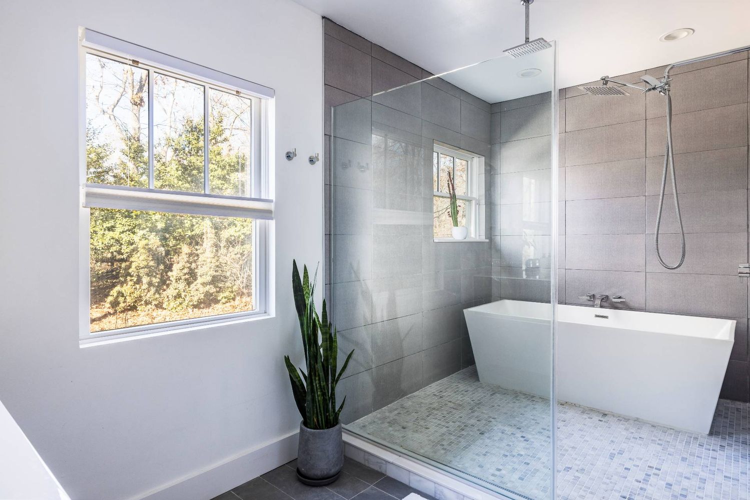 Modern bathroom with a freestanding tub, glass shower, gray tile, and a window overlooking trees.