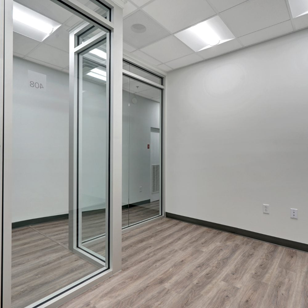 Empty office with glass partitions and gray wood-look flooring.