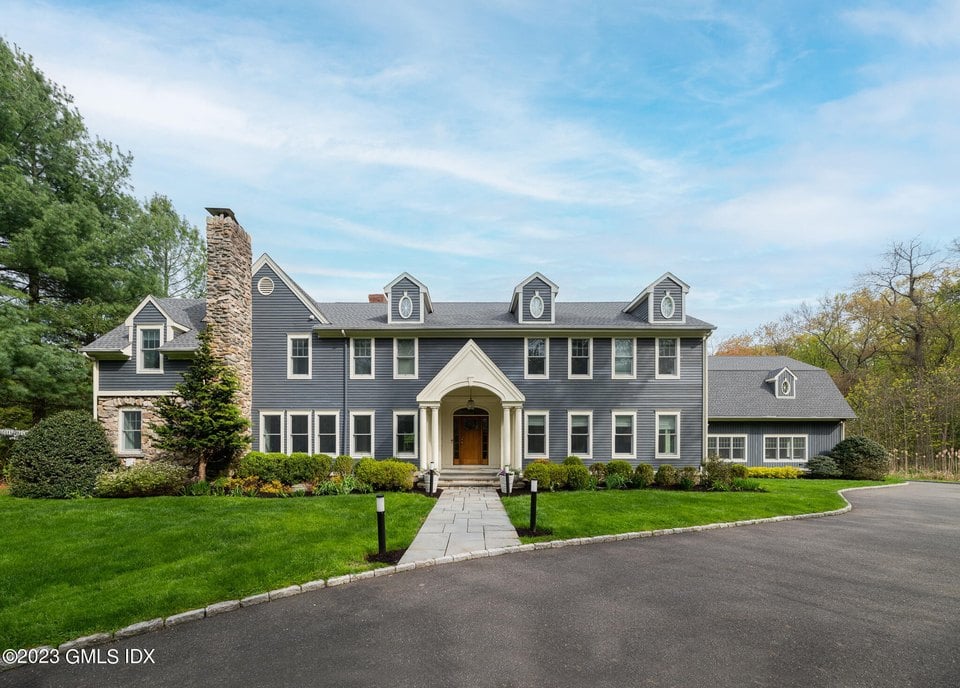 Gray-sided house with a tall stone chimney, three dormers, and a central covered entrance.