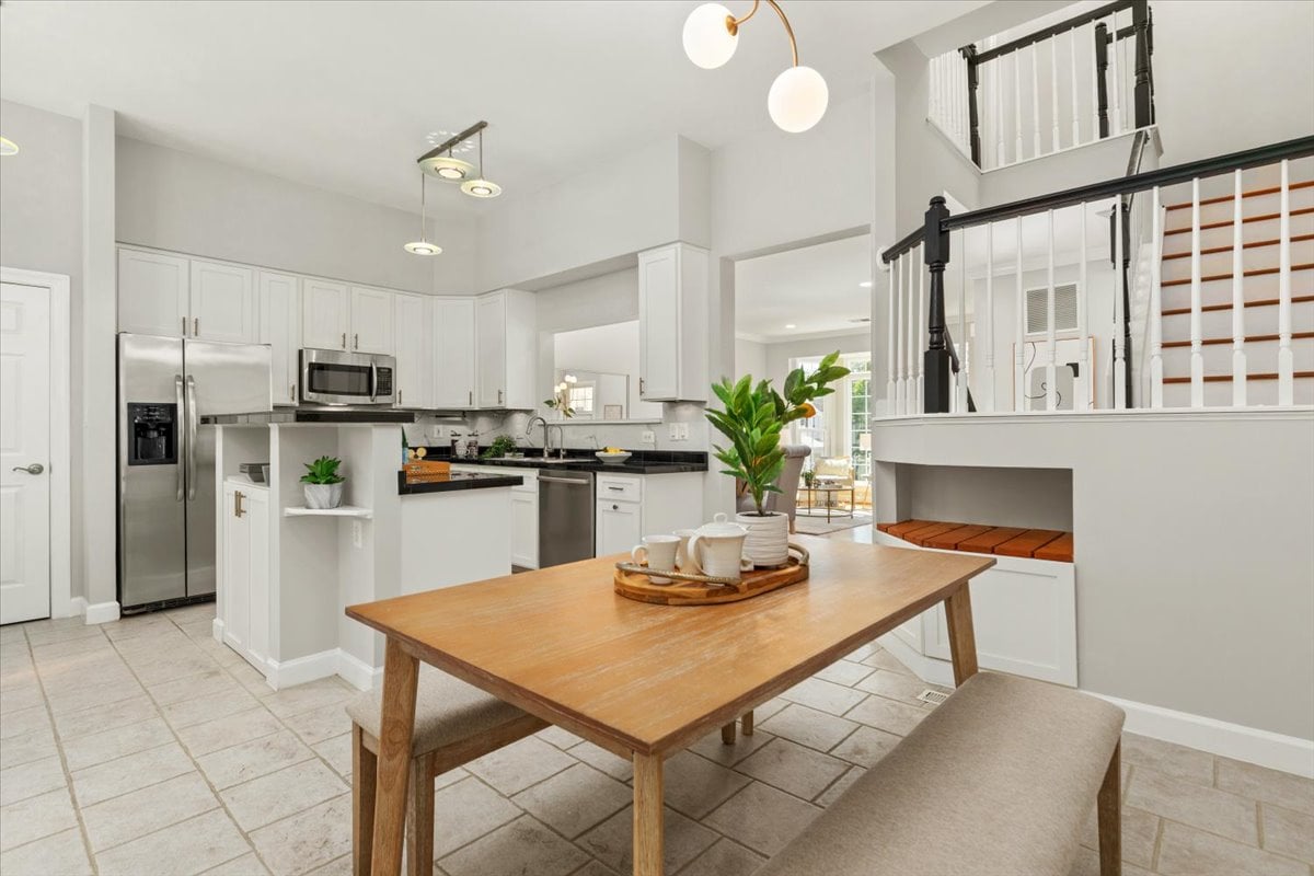 Open-plan kitchen with white cabinets, stainless appliances, and a wooden dining table beside a staircase and fireplace.