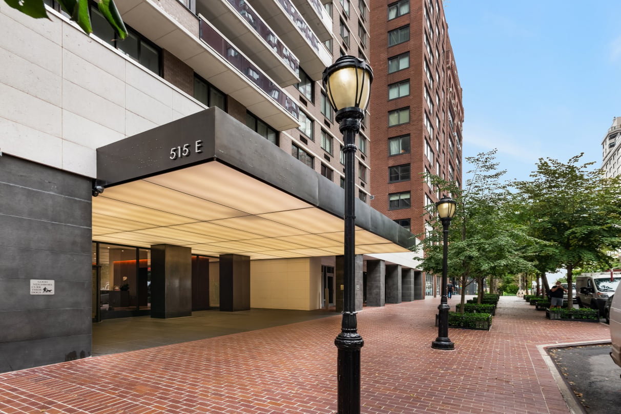 Condominium entrance beneath a broad illuminated canopy, beside a brick sidewalk and tree-lined street.