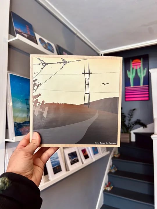 A hand holds a small square wooden art print depicting the Sutro Tower in San Francisco at sunset, with other artwork visible on shelves and a colorful cactus print on the wall.