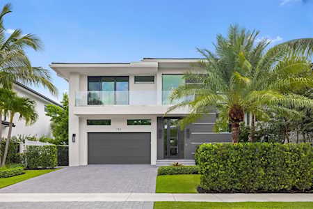 Modern two-story white home with a dark garage door, palm trees, and trimmed landscaping.
