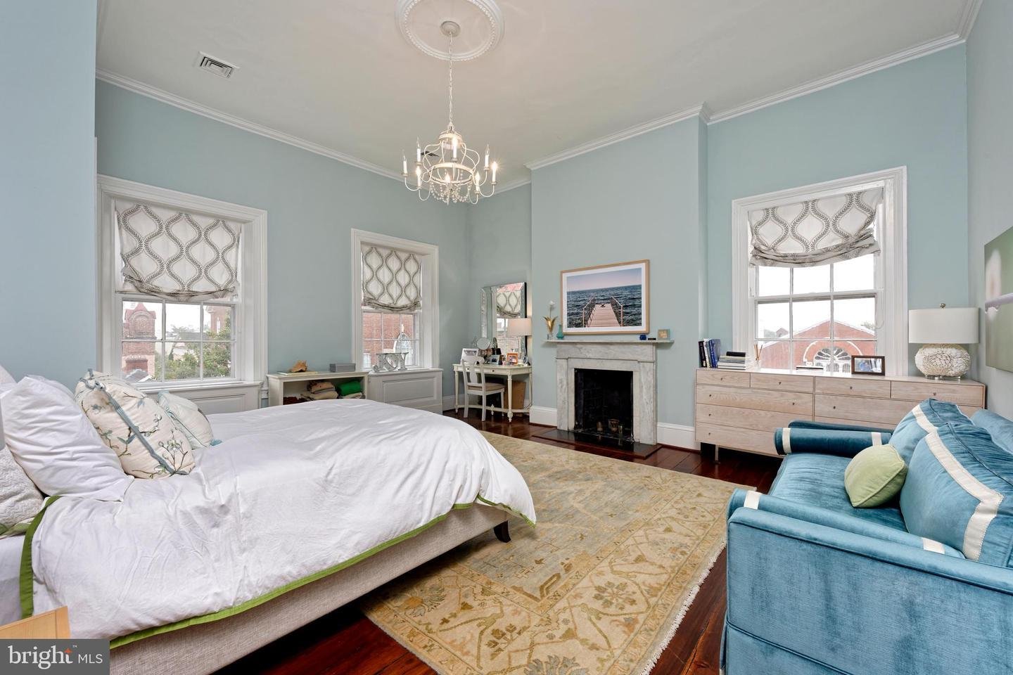 Furnished bedroom with pale blue walls, a fireplace, chandelier, and blue sofa beside a white bed.