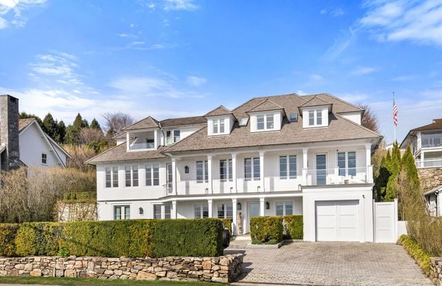Large white home with dormer windows, covered balconies, and an attached garage behind a hedge.